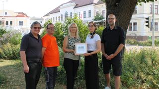 Fünf Personen stehen vor einem Gebäude, eine Frau hält ein gerahmtes Zertifikat. Sie alle lächeln und scheinen für ein Foto zu posieren.