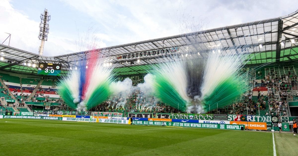 Ein Fußballstadion voller Fans, mit mehrfarbigem Rauch und Feuerwerk in der Luft und einem großen Banner mit Text und Logos.