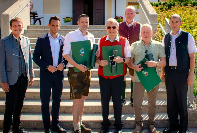 Fünf Männer stehen auf einer Treppe und halten grüne Ordner in der Hand, während sie für ein Foto posieren. Der Mann in der Mitte trägt eine Sonnenbrille und eine rote Weste. Der Mann auf der linken Seite trägt einen Anzug und Lederschuhe. Der Mann auf der rechten Seite trägt ein grünes Poloshirt und Khakihosen.