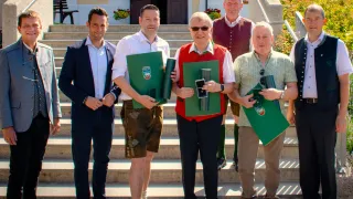 Fünf Männer stehen auf einer Treppe und halten grüne Ordner in der Hand, während sie für ein Foto posieren. Der Mann in der Mitte trägt eine Sonnenbrille und eine rote Weste. Der Mann auf der linken Seite trägt einen Anzug und Lederschuhe. Der Mann auf der rechten Seite trägt ein grünes Poloshirt und Khakihosen.