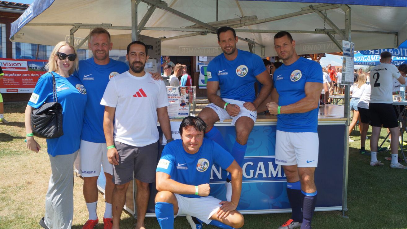 Fünf Männer in Sportuniformen, drei in Blau und zwei in Weiß, posieren für ein Foto unter einem Zelt. Ein Mann sitzt und die anderen stehen hinter ihm.