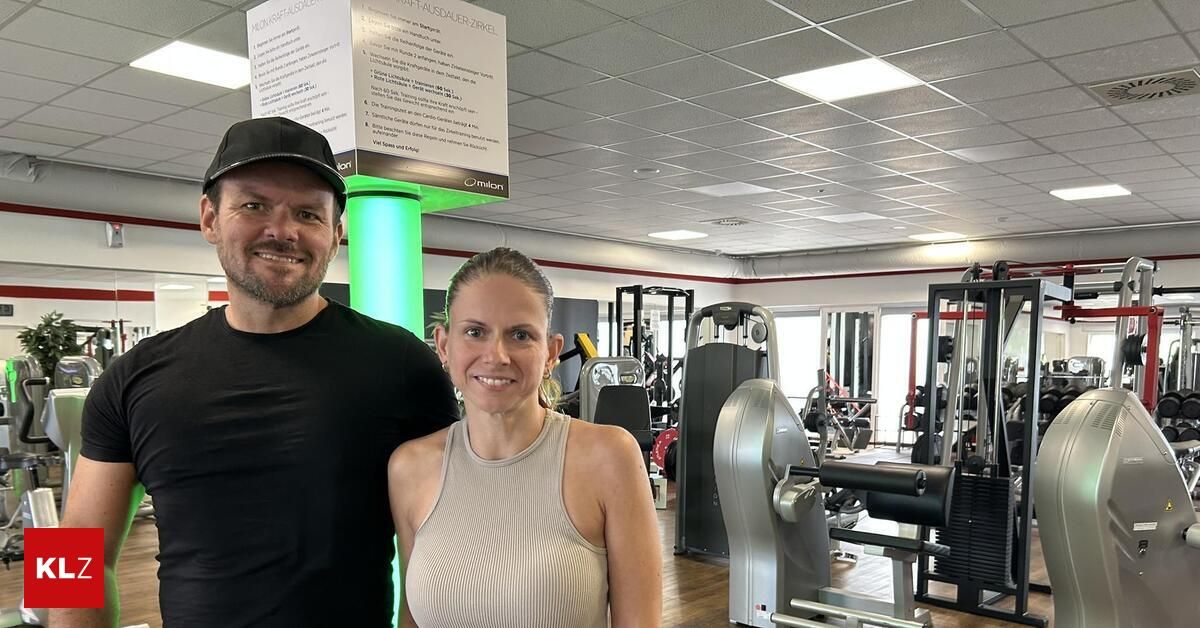 Ein Mann und eine Frau lächeln und posieren für ein Foto in einem Fitnessstudio. Der Mann trägt eine schwarze Kappe und ein schwarzes T-Shirt, und die Frau trägt ein ärmelloses Oberteil. Hinter ihnen steht eine grüne Säule mit einem Schild und verschiedenen Fitnessgeräten.