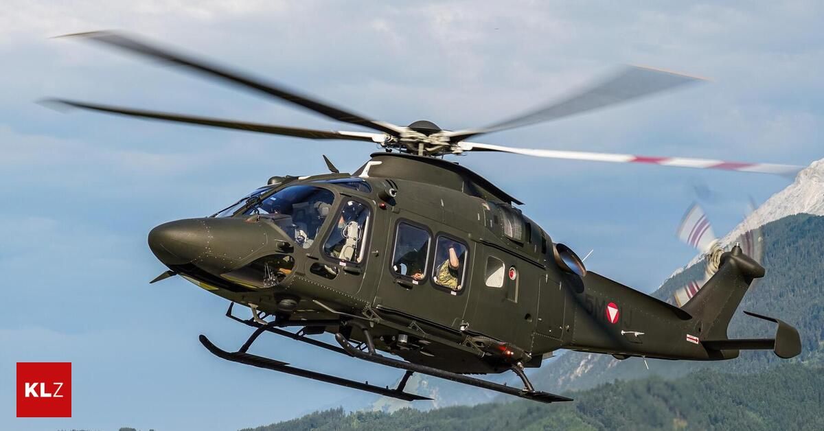Ein Militärhubschrauber mit einem roten Dreieckslogo an der Seite fliegt über einem bergigen Gelände.