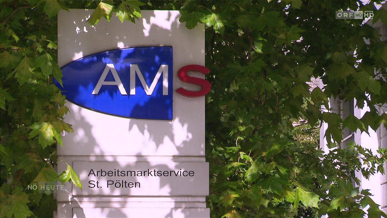 Ein weißer Schild mit blauen und roten Buchstaben steht vor einem Baum mit grünen Blättern und zeigt 'AMS' und 'Arbeitsmarktservice St. Pölten'.