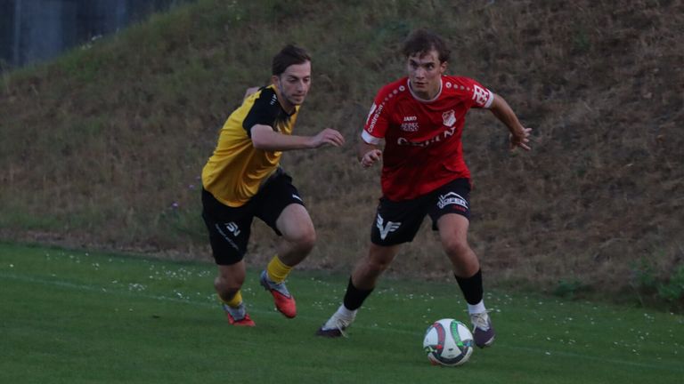 Zwei Fußballspieler konkurrieren auf einem Feld, einer in einem gelben und schwarzen Trikot, der andere in einem roten Trikot. Der Spieler in Rot läuft auf den Ball zu.