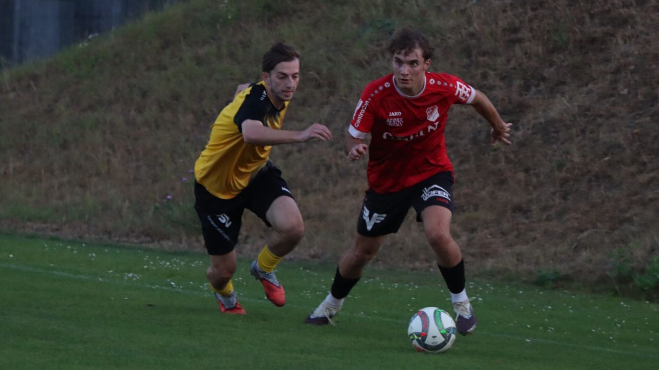 Zwei Fußballspieler konkurrieren auf einem Feld, einer in einem gelben und schwarzen Trikot, der andere in einem roten Trikot. Der Spieler in Rot läuft auf den Ball zu.