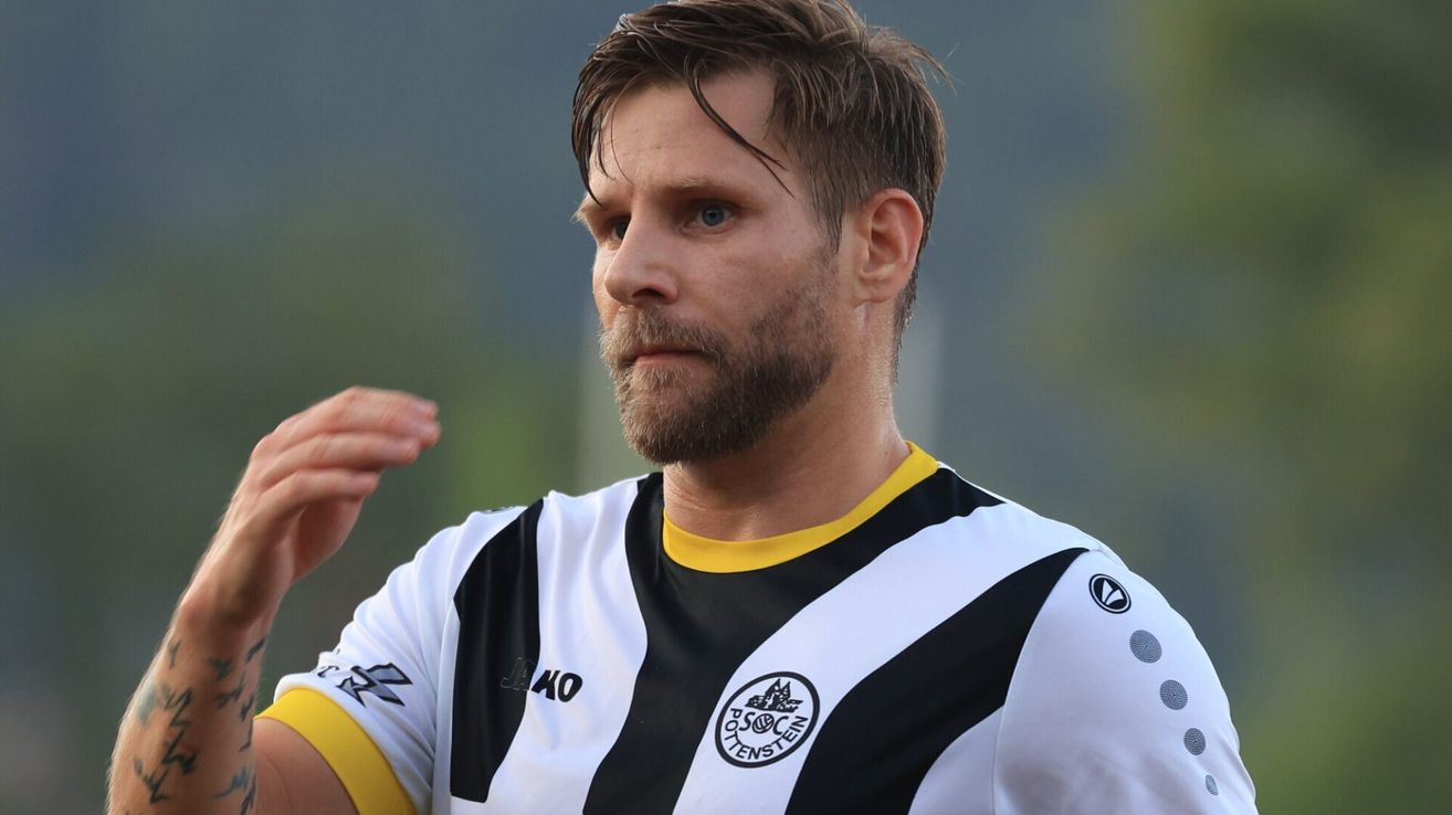 Nahaufnahme eines Mannes mit Bart, der ein schwarz-weiß gestreiftes Fußballtrikot mit gelbem Kragen trägt. Er schaut nach links, möglicherweise während eines Fußballspiels.