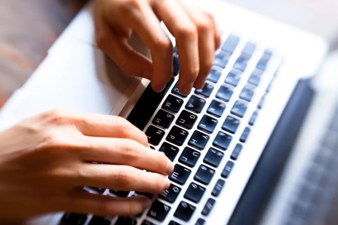 Eine Nahaufnahme der Hände einer Person, die auf einer Laptop-Tastatur mit schwarzen Tasten tippt.