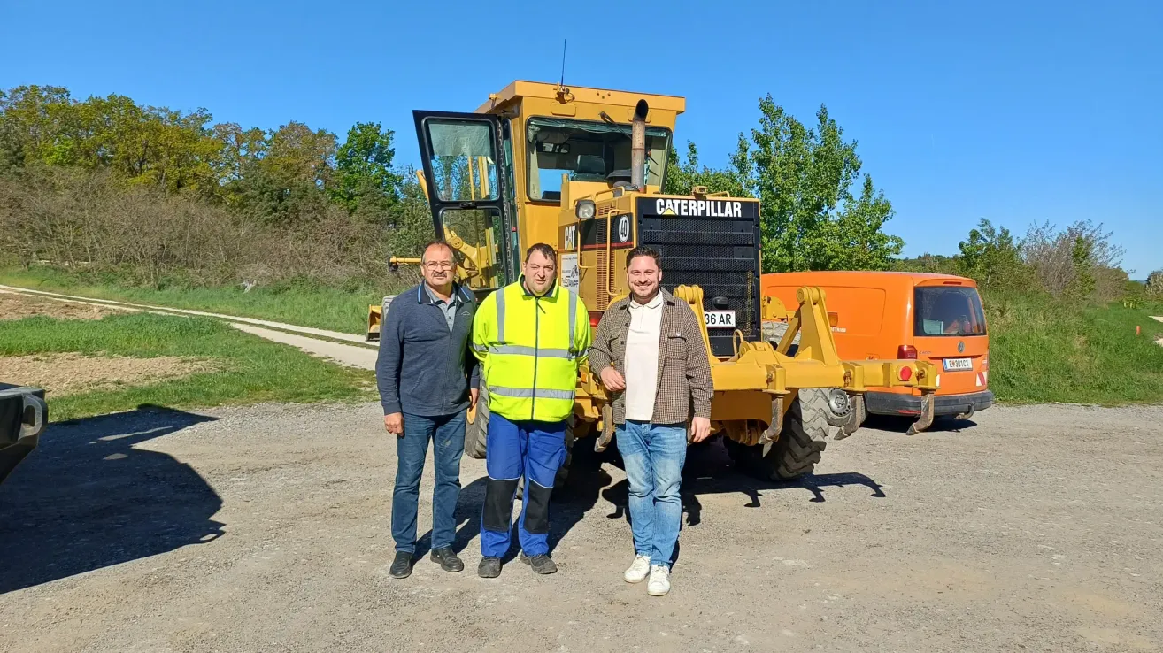 Drei Männer stehen vor einem gelben Caterpillar-Bagger und lächeln für die Kamera. Dahinter befindet sich eine Rasenfläche mit Bäumen und einem klaren blauen Himmel.