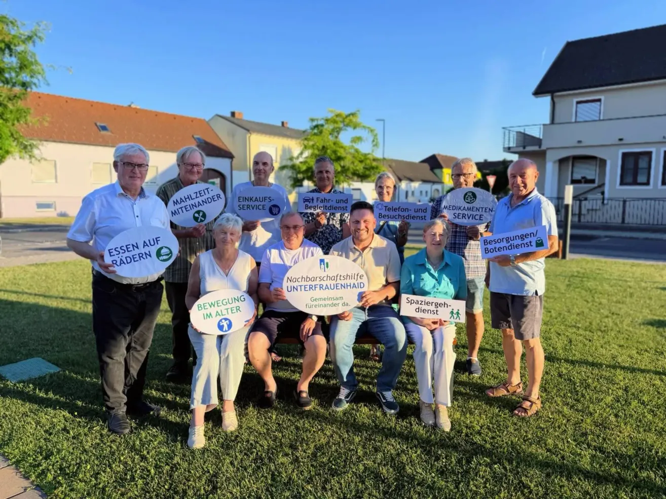 Eine Gruppe von Menschen steht in einem Park und hält Schilder mit den Aufschriften 'Nachbarschaftshilfe', 'Einkaufsservice' und 'Mobilität im Alter'. Sie lächeln und scheinen für ein Foto zu posieren.