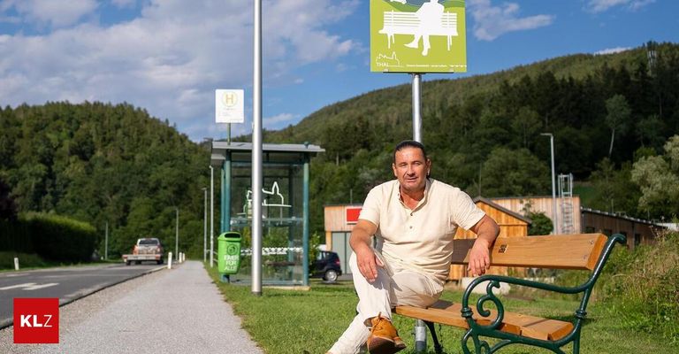 A man sits on a bench in a park with a sign behind him that reads 'Thal' and a trash bin labeled 'Eimer für alle'. He is wearing a white shirt and pants. In the background, there is a bus stop and a mountain.