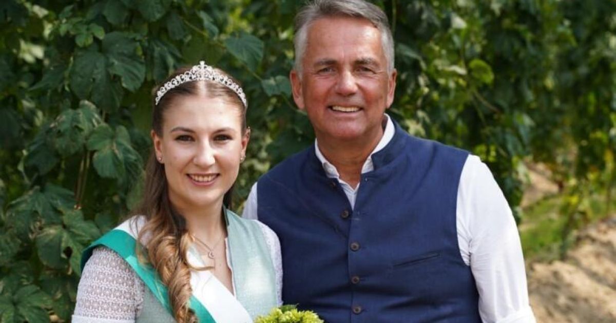 Ein Mann und eine Frau stehen nah beieinander und lächeln für ein Foto. Die Frau trägt eine Krone und eine Schärpe, während der Mann eine blaue Weste über einem weißen Hemd trägt. Sie stehen vor grünen Pflanzen.
