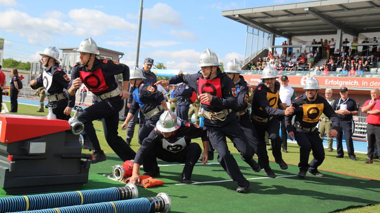 Eine Gruppe von Feuerwehrleuten in Uniform läuft auf einem Feld mit Feuerwehrschläuchen und Ausrüstung.
