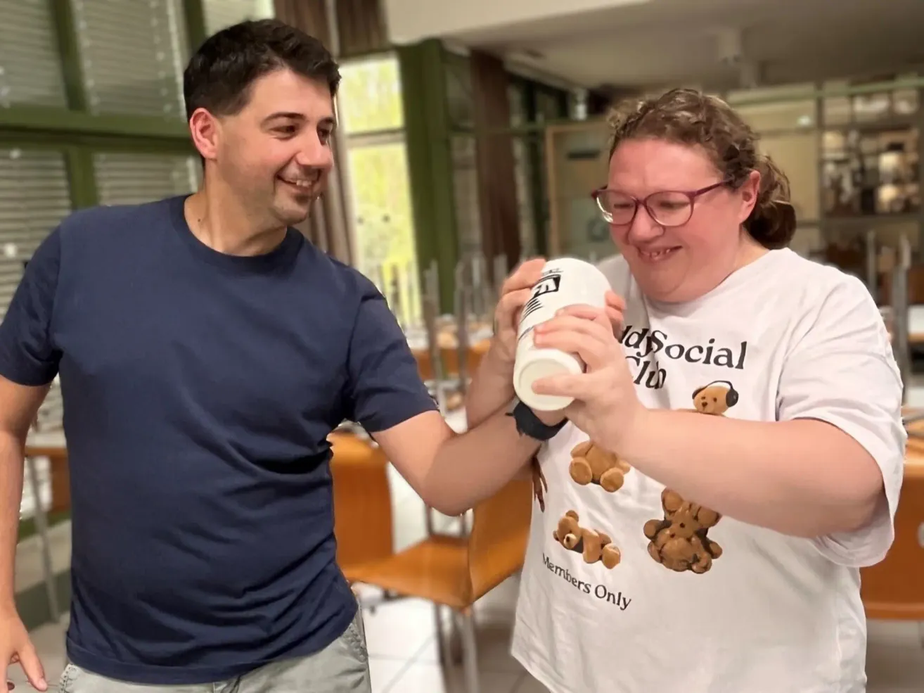 Ein Mann und eine Frau lächeln und halten einen Becher, möglicherweise bei einer sozialen Veranstaltung. Die Frau trägt ein weißes T-Shirt mit einem Bärendesign und den Worten 'Members Only'.