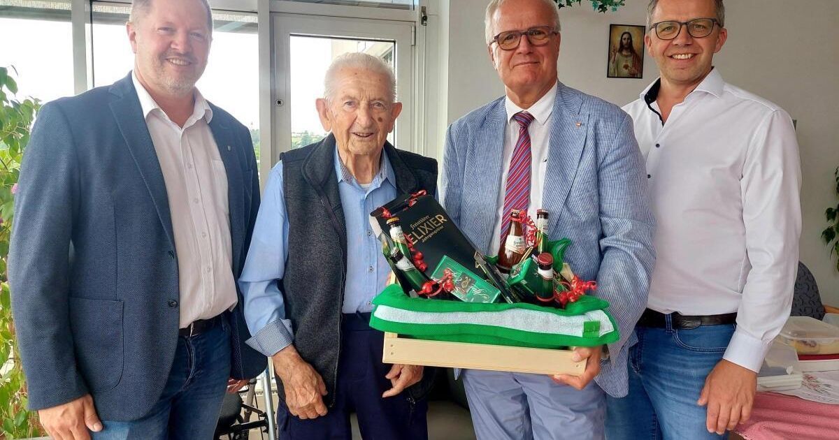 Vier Männer stehen drinnen zusammen, einer hält einen Geschenkkorb mit Flaschen und einem Buch. Sie lächeln und scheinen einen Anlass zu feiern.