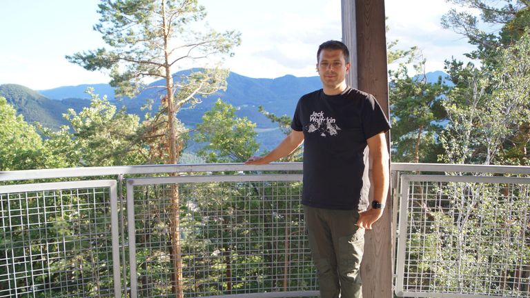 Ein Mann in einem schwarzen Jack Wolfskin-Shirt steht auf einem Balkon mit einer Metallbrüstung und blickt auf eine bewaldete Berglandschaft.