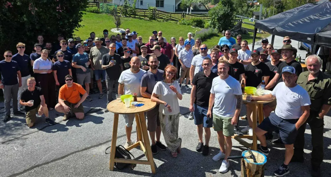 Eine Gruppe von Menschen, hauptsächlich Männer, versammelt sich im Freien. Einige stehen, andere sitzen. Sie alle lächeln und scheinen für ein Foto zu posieren. Es gibt Holztische und Stühle im Bereich. Im Hintergrund befinden sich Häuser und ein Zaun.