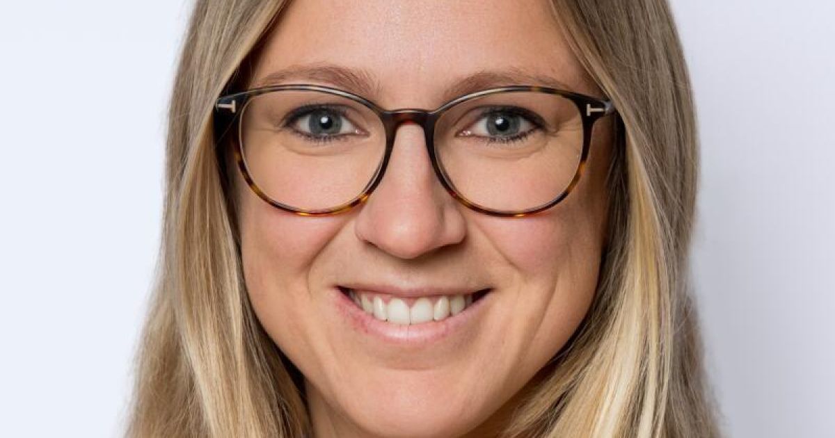 Eine Frau mit blonden Haaren, die eine Brille trägt und für die Kamera lächelt. Sie hat helle Haut und blaue Augen.