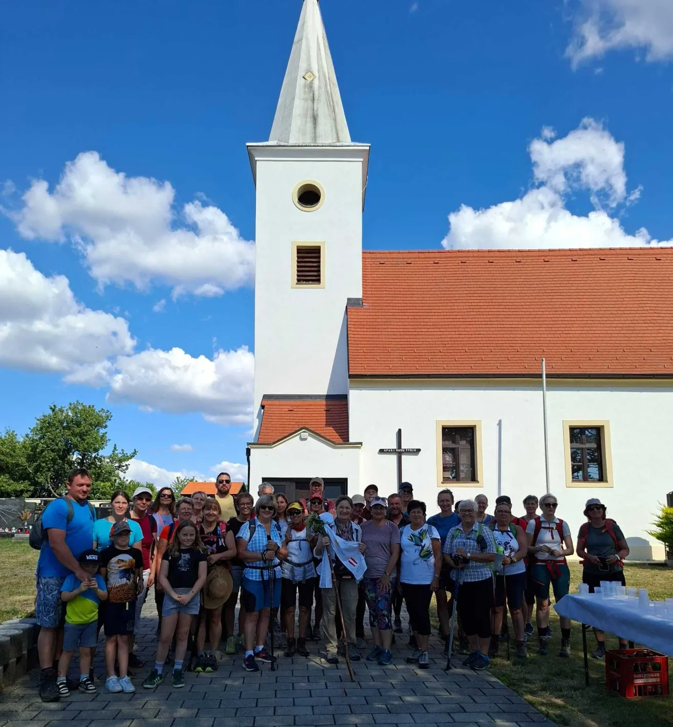 Eine Gruppe von Menschen vor einer Kirche mit rotem Dach und weißem Turm, unter einem blauen Himmel mit Wolken.
