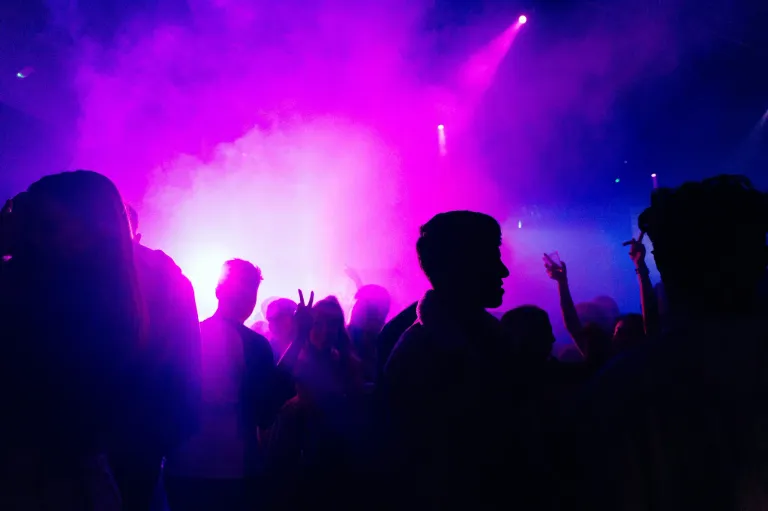 Eine Menschenmenge tanzt in einem Club, während lila und pinke Lichter die Szene erhellen.