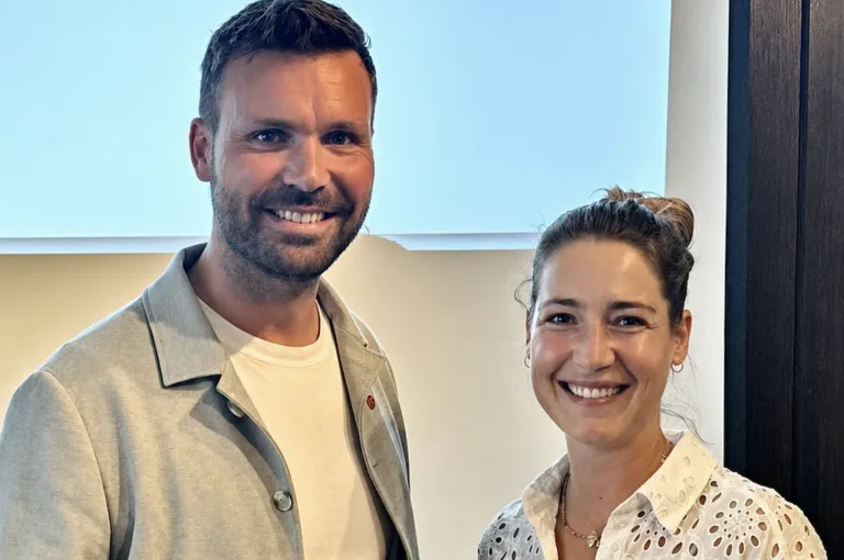 Ein Mann und eine Frau stehen eng beieinander und lächeln beide hell für ein Foto. Der Mann hat einen Bart und trägt ein helles Hemd und eine Jacke, während die Frau ihr Haar zu einem Dutt gebunden hat und ein weißes Blusenhemd trägt. Sie scheinen sich in einem gut beleuchteten Raum mit einem hellblauen Hintergrund zu befinden.