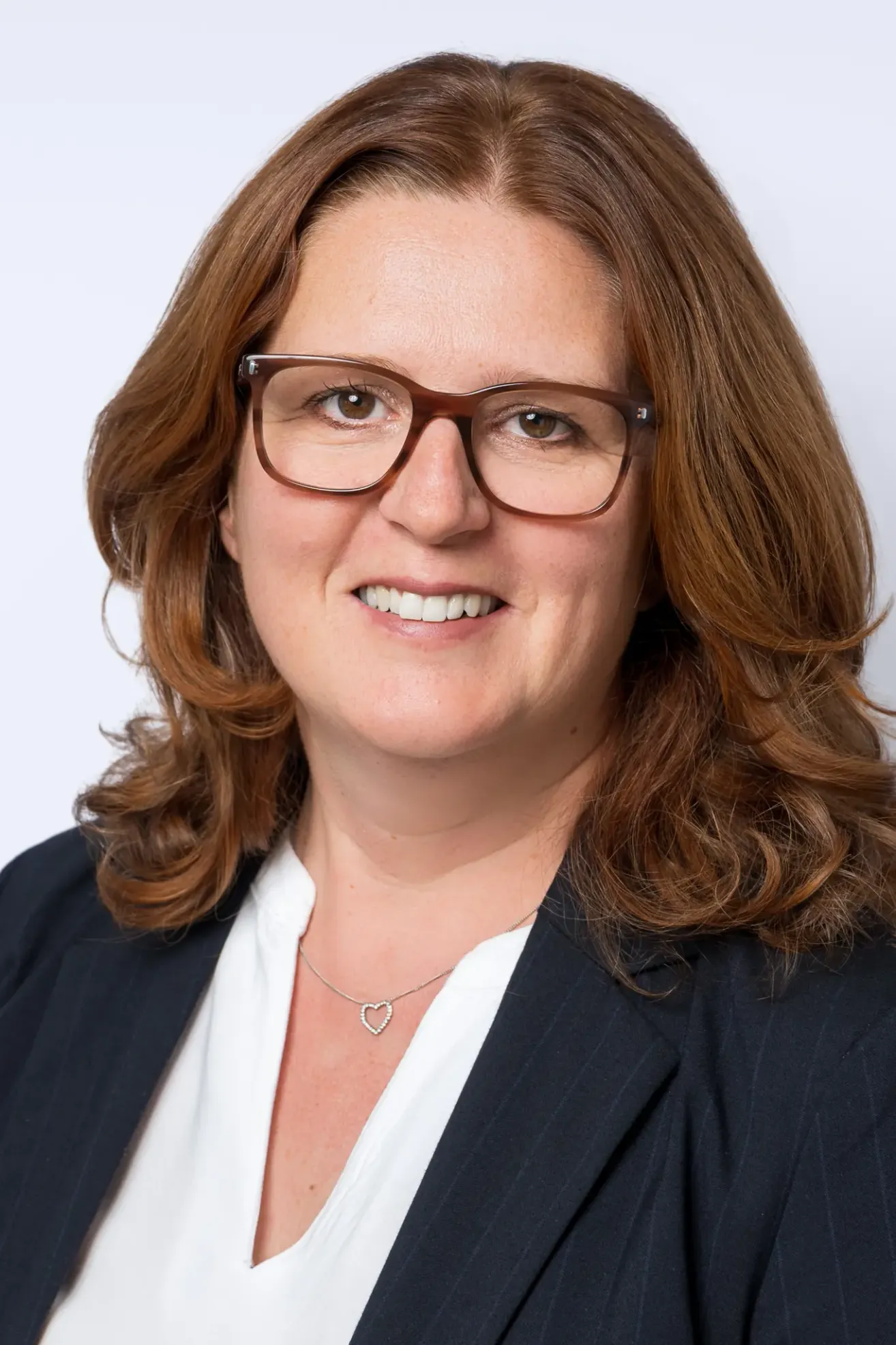 Eine Frau mit braunen Haaren, die eine Brille und einen schwarzen Blazer trägt, lächelt und sieht in die Kamera. Sie hat ein weißes Hemd und eine Herzanhänger-Halskette.