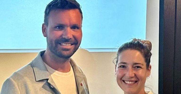 Ein Mann und eine Frau lächeln und stehen eng nebeneinander vor einem Fenster.