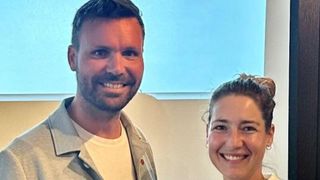 Ein Mann und eine Frau lächeln und stehen eng nebeneinander vor einem Fenster.