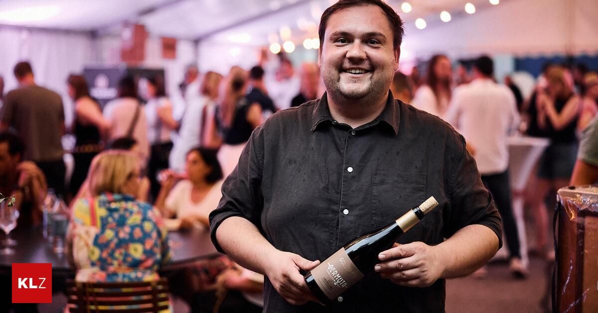 Ein Mann in einem schwarzen Hemd hält eine Flasche Wein bei einer Veranstaltung mit Menschen im Hintergrund.