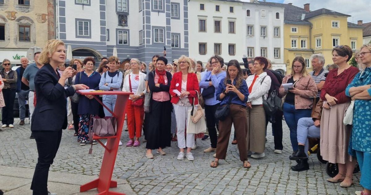 Eine Gruppe von Menschen steht auf einem kopfsteingepflasterten Platz, einige tragen Brillen und halten Telefone, mit einem roten Ständer im Vordergrund. Hinter ihnen sind Gebäude mit vielen Fenstern zu sehen.