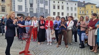 Eine Gruppe von Menschen steht auf einem kopfsteingepflasterten Platz, einige tragen Brillen und halten Telefone, mit einem roten Ständer im Vordergrund. Hinter ihnen sind Gebäude mit vielen Fenstern zu sehen.