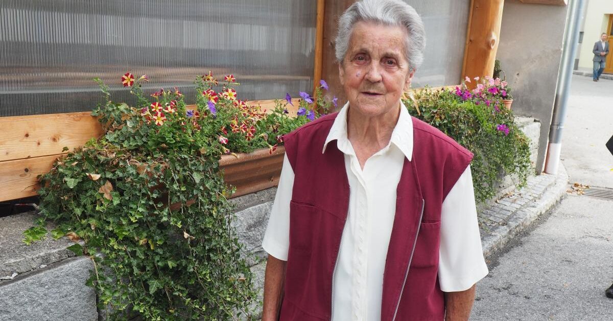 Eine ältere Frau mit weißen Haaren steht vor einem Fenster mit Blick auf bunte Blumen und Grün. Sie trägt ein weißes Hemd und eine weinrote Weste.