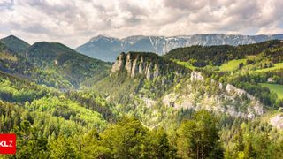 Ein Panoramablick auf eine Bergregion mit einer Brücke im Tal, umgeben von üppigem Grün und einer bewölkten Himmelslandschaft.