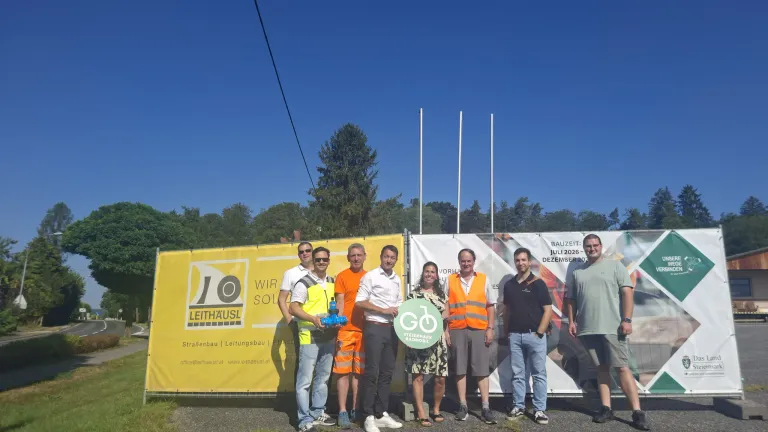 Eine Gruppe von Personen in Warnwesten und Turnschuhen steht vor einem gelben Banner mit den Texten 'Leithaeusl' und 'Stiermark Radmobil'. Sie halten ein grünes Schild mit einem Fahrrad darauf. Hinter ihnen sind Bäume und ein klarer blauer Himmel.
