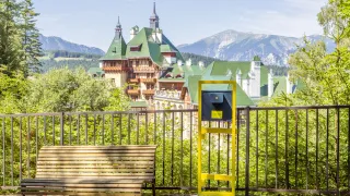 Eine Holzbank und ein gelber Ticketschalter stehen vor einem großen, grün gedeckten Gebäude mit Berghintergrund.