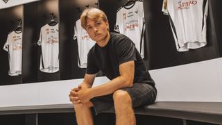 Ein Mann sitzt auf einer Bank in einem Umkleideraum mit mehreren Trikots, die hinter ihm hängen. Er trägt ein schwarzes Shirt und Shorts.