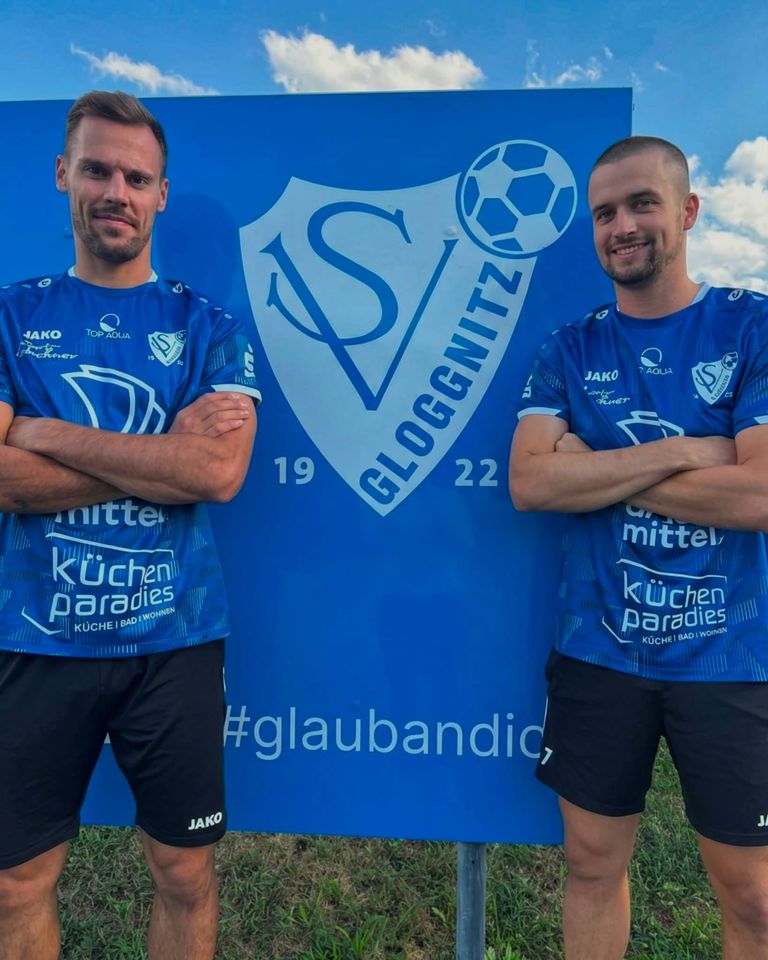 Zwei Männer in blauen Trikots mit dem Sponsorenlogo 'Kuchenparadies' stehen vor einem blauen Banner mit einem Fußball und dem Text 'VSGLOGGNITZ'.