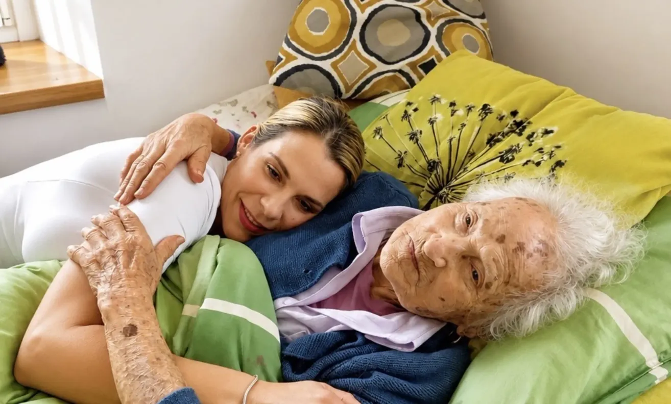Eine ältere Frau und eine jüngere Frau liegen im Bett, die jüngere Frau lächelt und hält die Hand der älteren Frau.