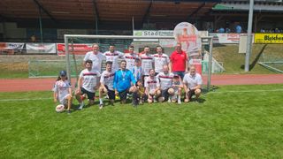 Eine Fußballmannschaft posiert für ein Foto vor einem Tor mit einem Banner dahinter. Sie tragen alle weiße Hemden und Shorts, einige mit Medaillen um den Hals.