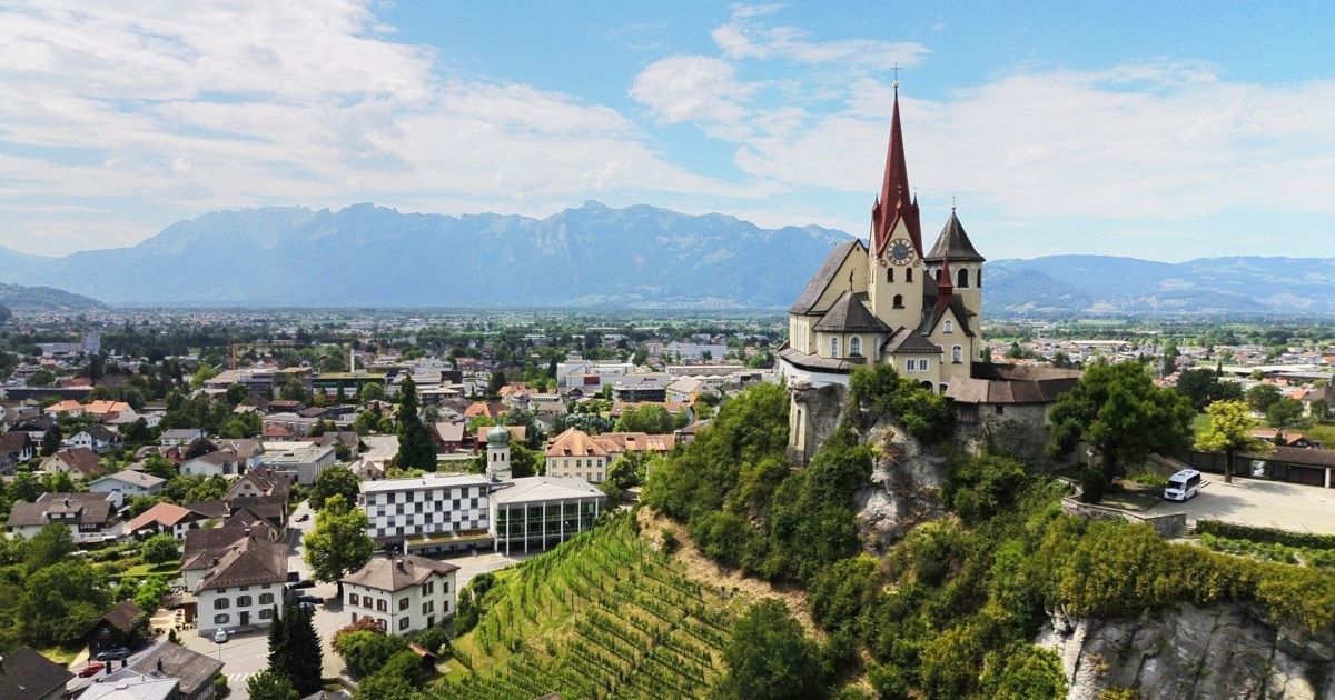 Luftaufnahme einer Stadt mit einer Kirche auf einem Hügel, umgeben von Bergen und Weinbergen.