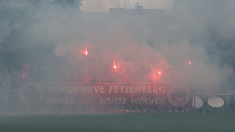 Eine Gruppe von Menschen, die rote Fackeln in einer nebligen Atmosphäre halten, stehen hinter einem Banner, auf dem 'Nordkurve FetzenBerg' und 'White Wolves' steht.