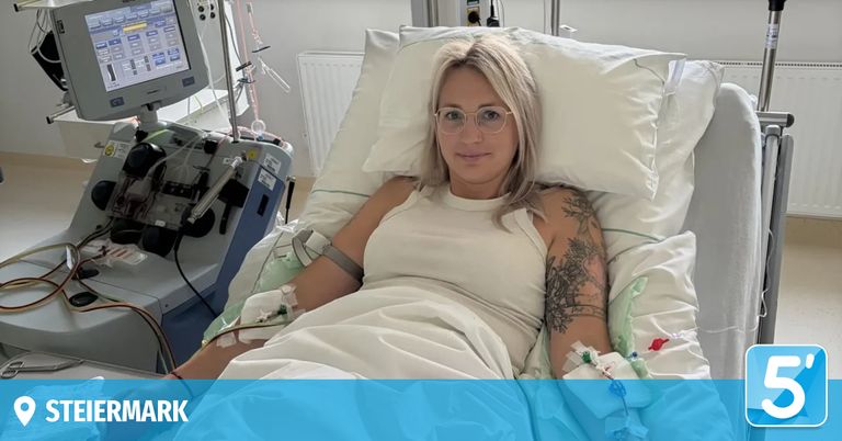 Eine Frau mit blonden Haaren liegt in einem Krankenhausbett, trägt eine Brille und ein ärmelloses Kleid. Sie hat eine Tätowierung auf ihrem rechten Arm und medizinische Geräte sind an ihr befestigt.