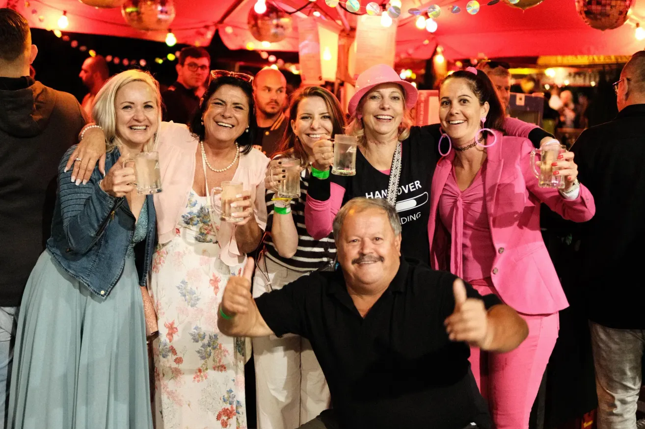 Eine Gruppe lächelnder Erwachsene, darunter Frauen in rosa und weißen Outfits und ein Mann in Schwarz, posieren für ein Foto bei einer Party mit Getränken und Disco-Lichtern.