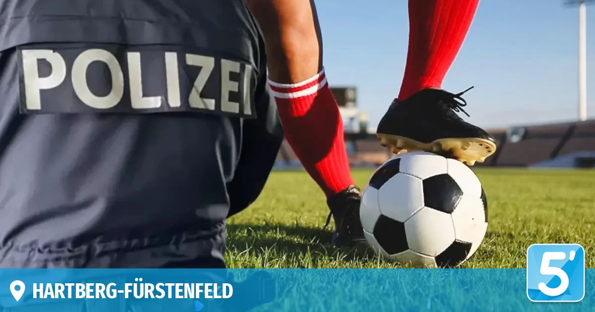 Ein Fußballspieler in roten Socken und schwarzen Stollenschuhen steht kurz davor, einen Fußball auf einem grünen Feld zu schießen, während ein Polizeibeamter in einer schwarzen Uniform zusieht.