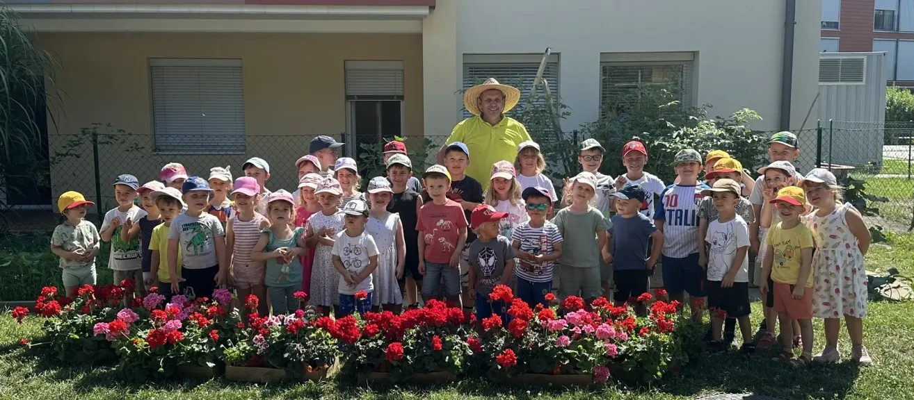 Eine Gruppe von Kindern und einem Mann in einem gelben Shirt und Strohhut stehen vor einem Gebäude mit roten Blumen im Vordergrund.