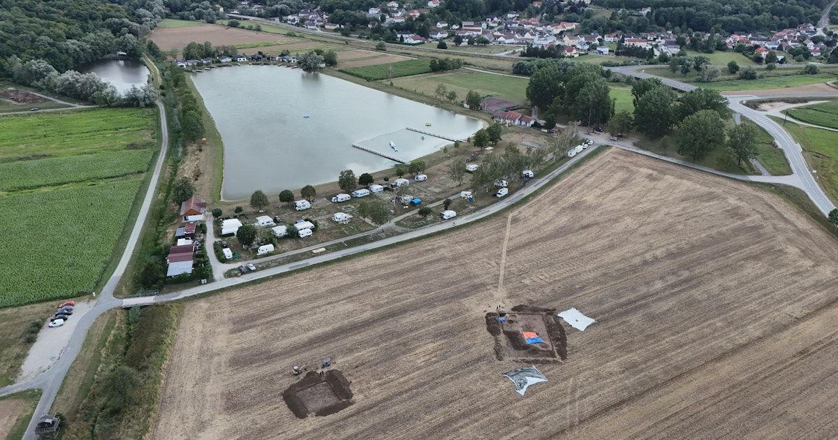 An aerial view of a lake with several boats docked on the shore, surrounded by a large field with parked vehicles and excavation sites.