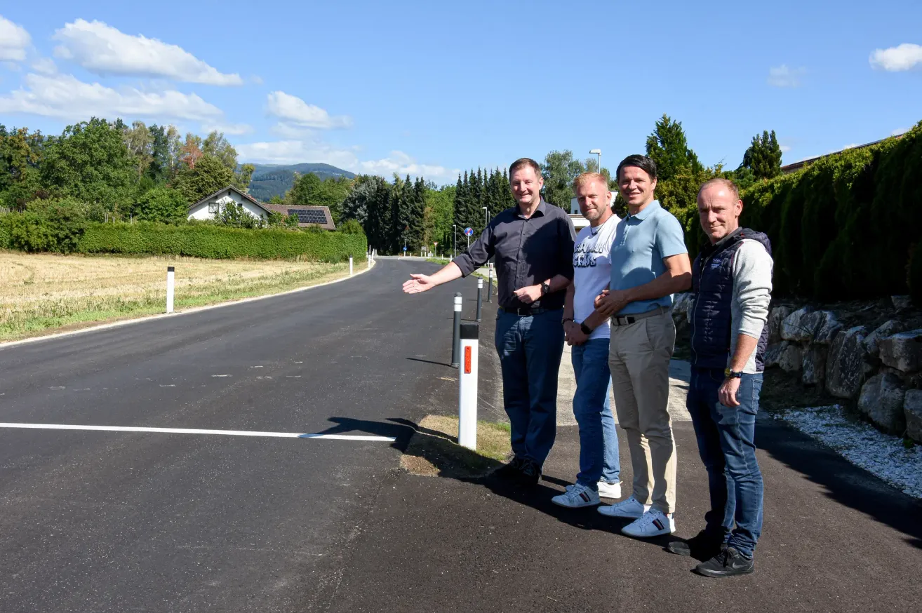 Vier Männer stehen auf einer neuen Straße, lächeln und posieren für ein Foto. Sie sind von viel Grün umgeben, mit Häusern und Bergen im Hintergrund.