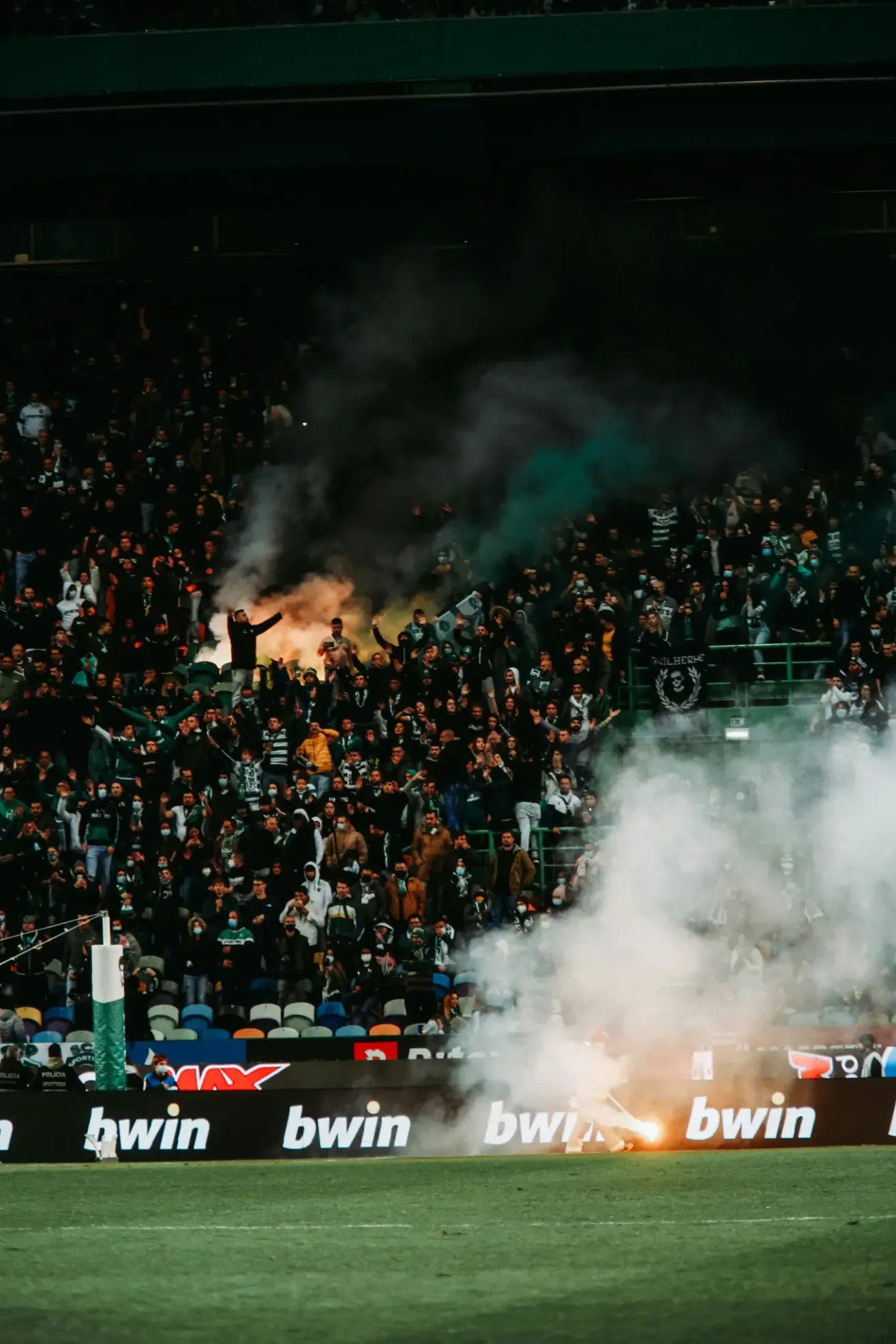 Eine Menschenmenge bei einer Sportveranstaltung, einige halten Fahnen, mit Rauch und Feuer im Vordergrund.