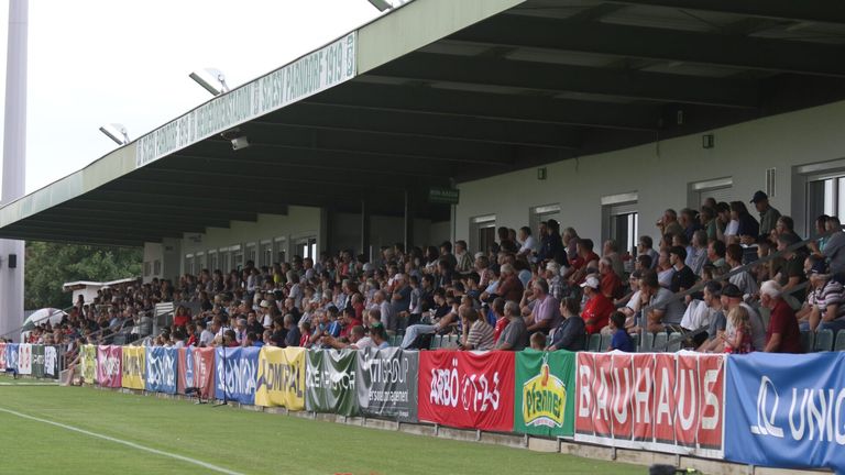 Eine große Menschenmenge sitzt in den Tribünen eines Stadions und schaut sich ein Spiel an. Die Tribünen sind mit bunten Bannern und Flaggen geschmückt. Das Spielfeld ist grün und gut gepflegt.