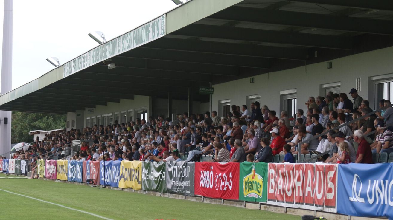 Eine große Menschenmenge sitzt in den Tribünen eines Stadions und schaut sich ein Spiel an. Die Tribünen sind mit bunten Bannern und Flaggen geschmückt. Das Spielfeld ist grün und gut gepflegt.